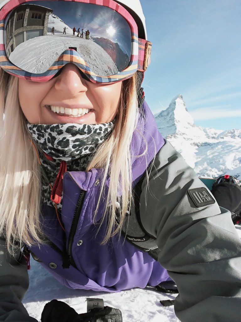 Woman in snowboard gear, taking a selfie with skiers in her goggle reflection.