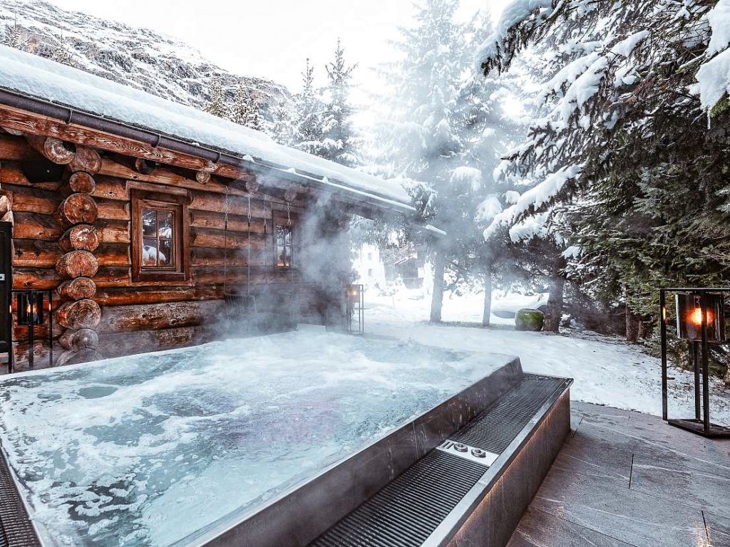 A steaming hot tub surrounded by snow and a wooden chalet at the Alpenhof Zermatt.