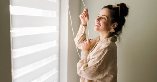 Happy woman enjoying morning sunlight through blinds.