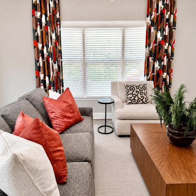 Bright living room with gray sofa, white armchair, colorful curtains, and wooden coffee table.
