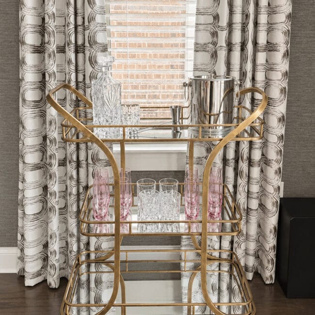 Gold bar cart with glassware and decanters in front of window with patterned curtains.