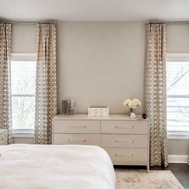 Bright bedroom with large windows, beige curtains, and white furniture.