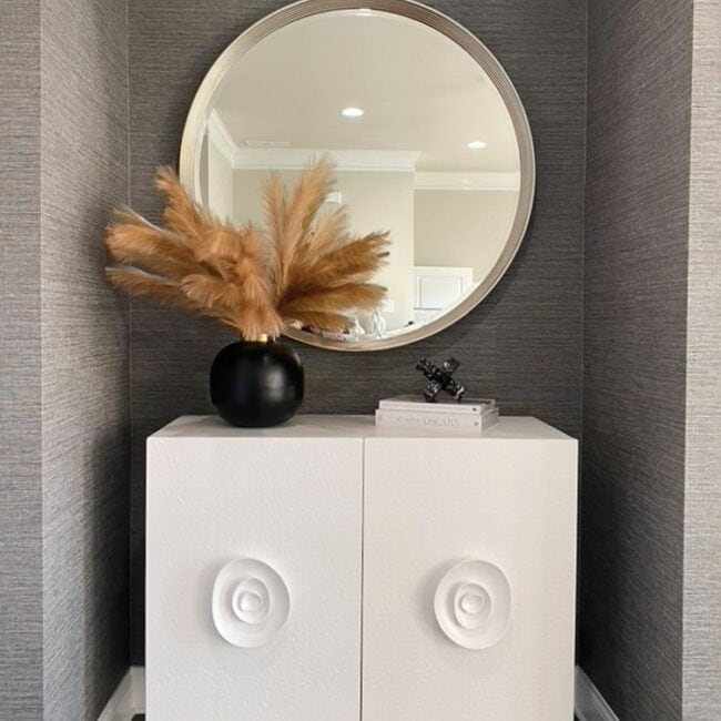Round mirror above white cabinet with black vase and pampas grass.