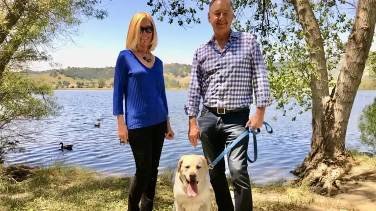 A vibrant dog park bathed in sunlight features abundant greenery with mature trees, offering ample shade for leisurely outdoor activities. In the distance is a well-equipped playground that presents an additional activity area. An inviting picnic table spreads out nearby, ideal for enjoying meals al fresco or taking relaxing breaks from play. A neatly-placed trash bin aids in maintaining the park's cleanliness for everyone's enjoyment. Dominating the scene is a generously wide dirt path gracefully curving through the expanse of grass, promising exploratory walks and lively fetch games.