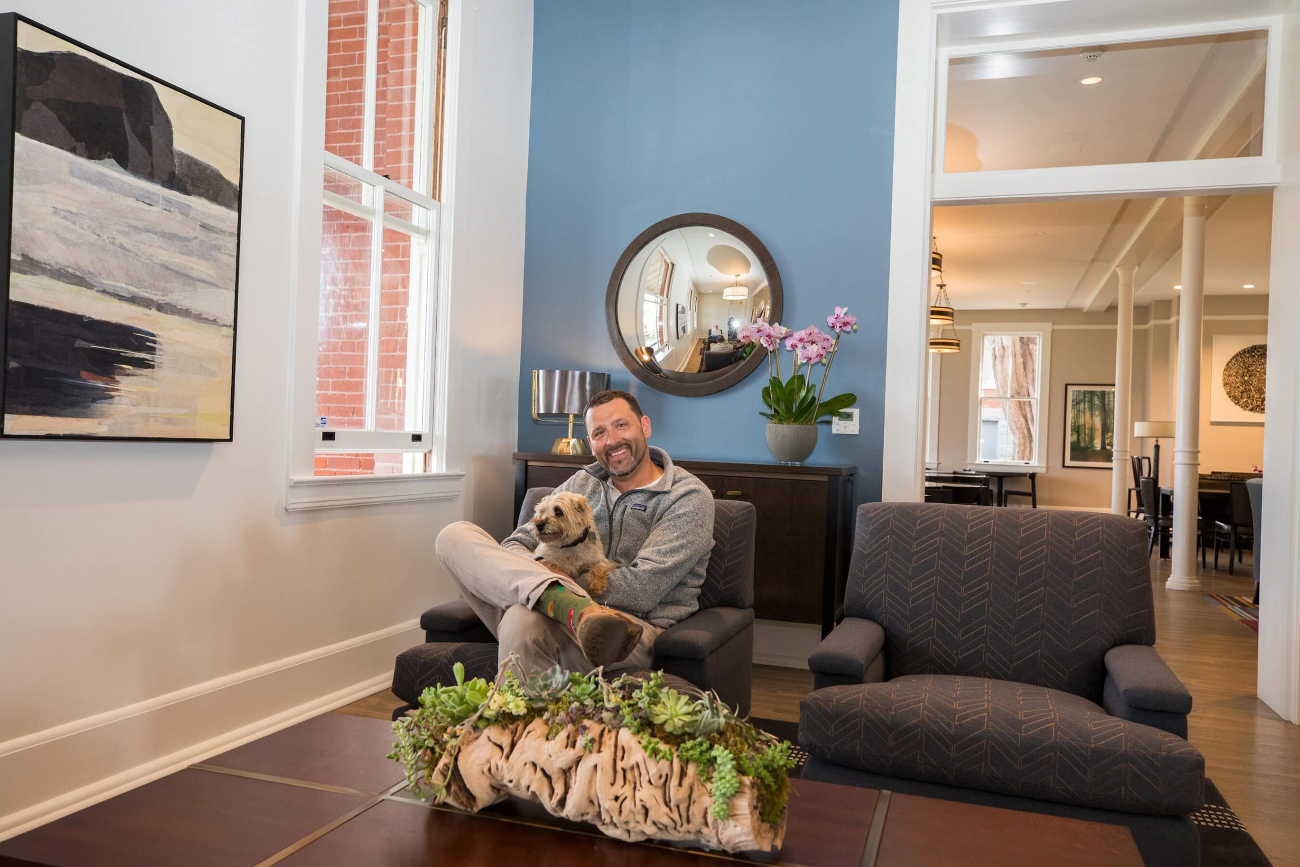 A vibrant dog park bathed in sunlight features abundant greenery with mature trees, offering ample shade for leisurely outdoor activities. In the distance is a well-equipped playground that presents an additional activity area. An inviting picnic table spreads out nearby, ideal for enjoying meals al fresco or taking relaxing breaks from play. A neatly-placed trash bin aids in maintaining the park's cleanliness for everyone's enjoyment. Dominating the scene is a generously wide dirt path gracefully curving through the expanse of grass, promising exploratory walks and lively fetch games.