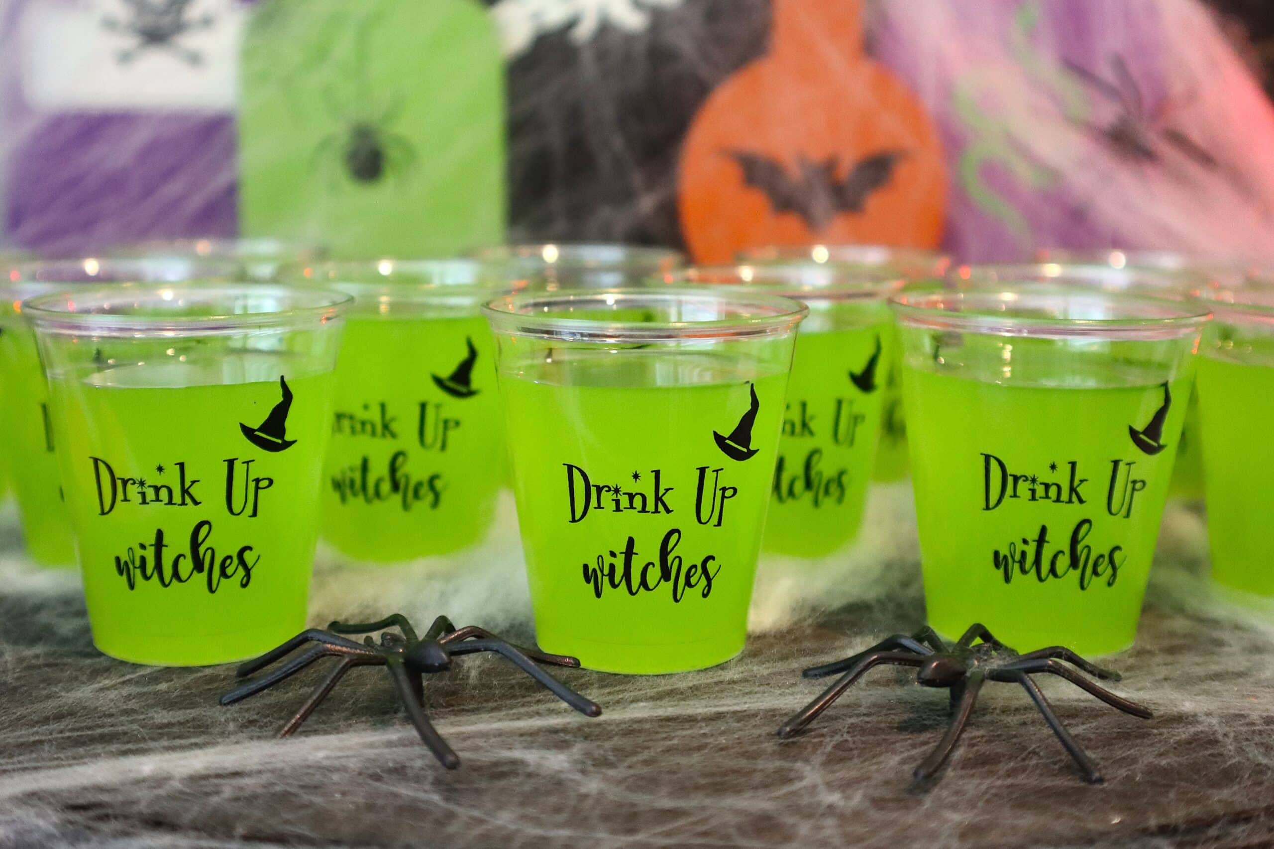 Green Halloween shot glasses with 'Drink Up Witches' text and spider decorations.
