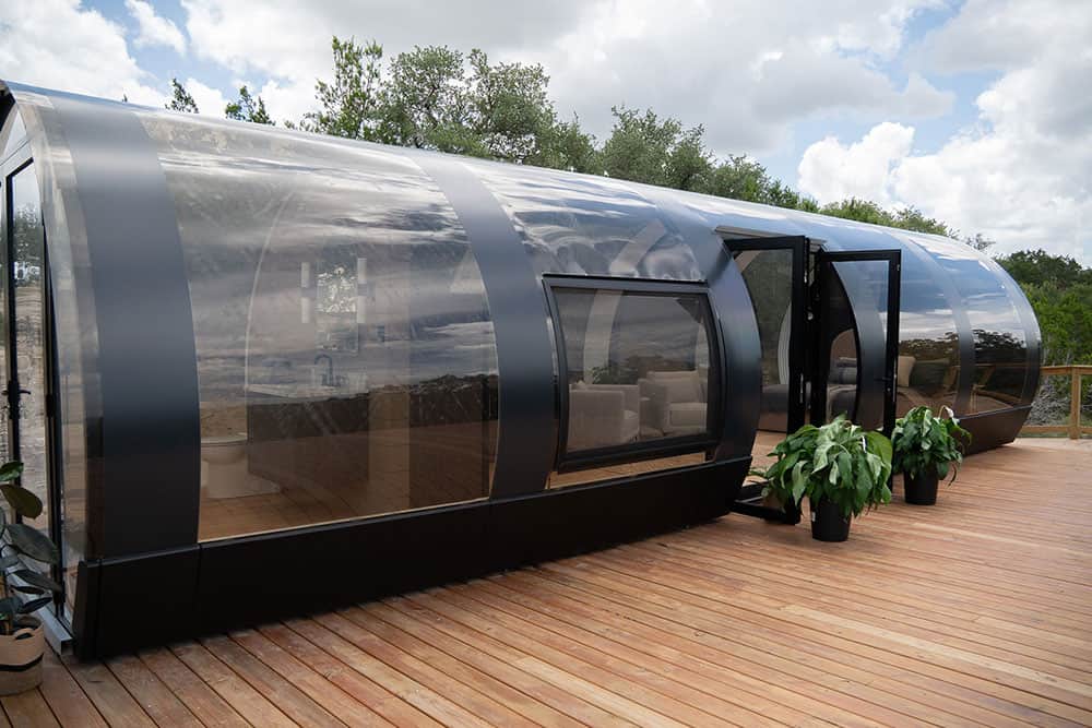 Sleek, modern glass outdoor living pod with black metal accents, expansive windows, and wooden deck, ideal for luxury residential or hospitality use.