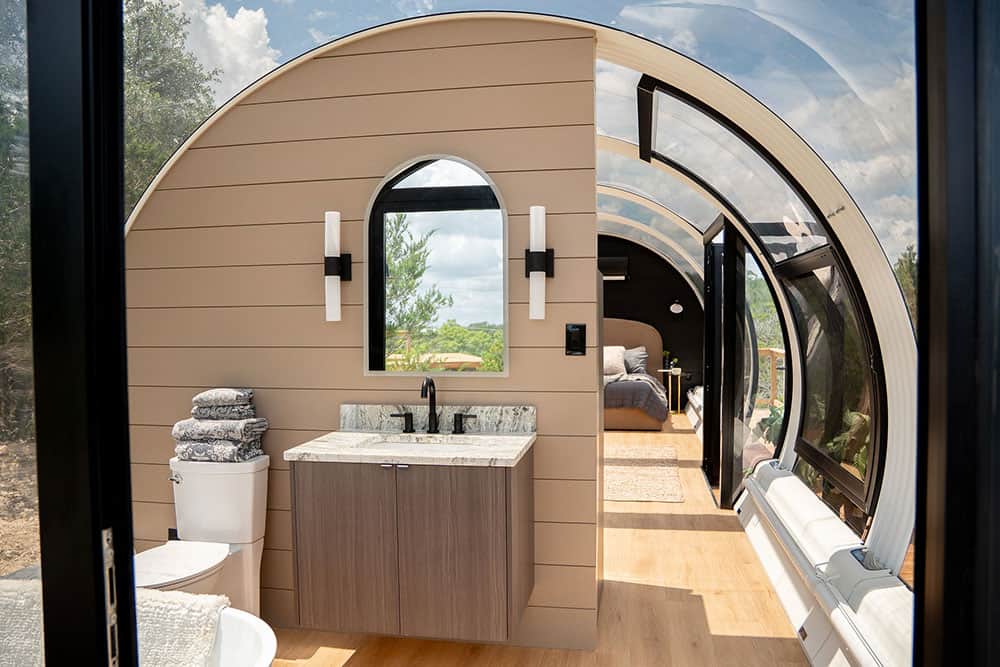 Modern tiny house with arched glass walls and interior bathroom featuring a vanity, toilet, and towels, showcasing innovative space-saving design solutions.