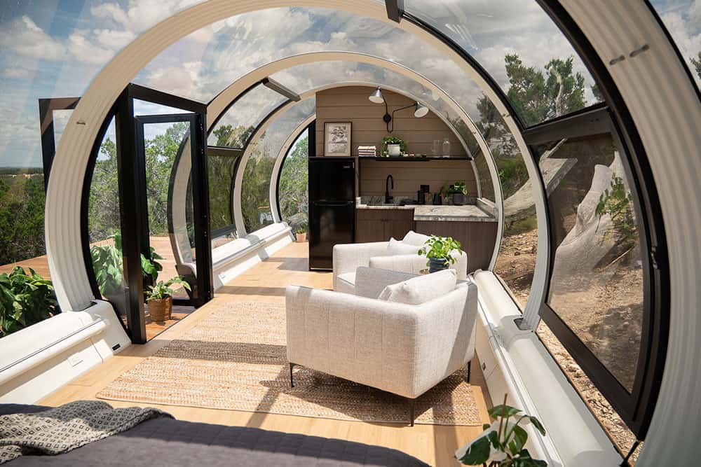 Modern glass-enclosed tiny home with open kitchen, cozy seating area, and panoramic views, showcasing innovative compact living solutions by EJH Distribution.