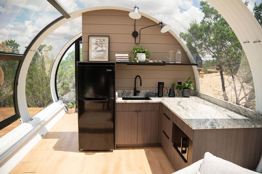 Compact modern kitchenette inside a stylish, eco-friendly tiny home with large arched windows, green outdoor scenery, and minimalistic design, perfect for sustainable living and urban retreats.