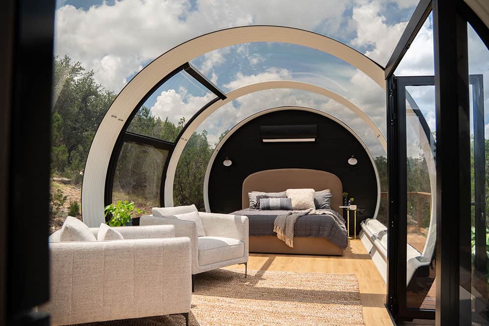 Modern glass-enclosed bedroom with curved architecture and scenic views, featuring contemporary furniture and a cozy bed, ideal for luxury interior design and innovative architectural projects.