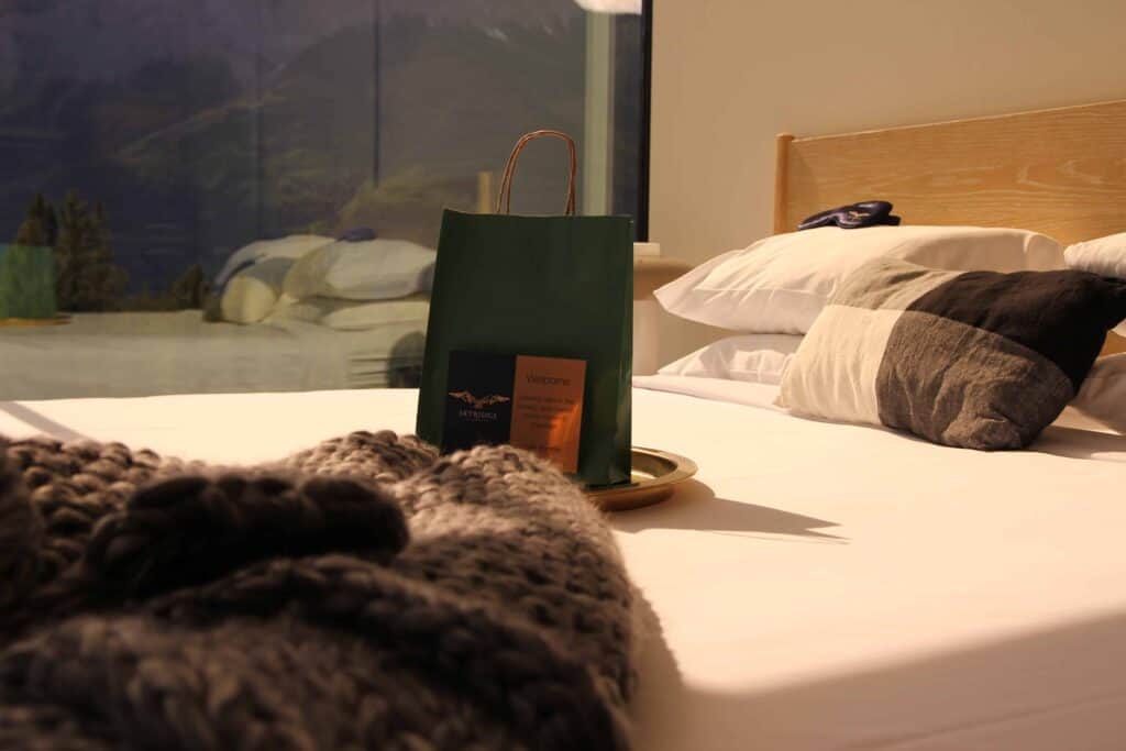 Deep hotel bed with a cozy knitted blanket, a welcome gift bag on a tray, and a view of mountains through a large window, showcasing comfort and hospitality at EJH Distribution properties.