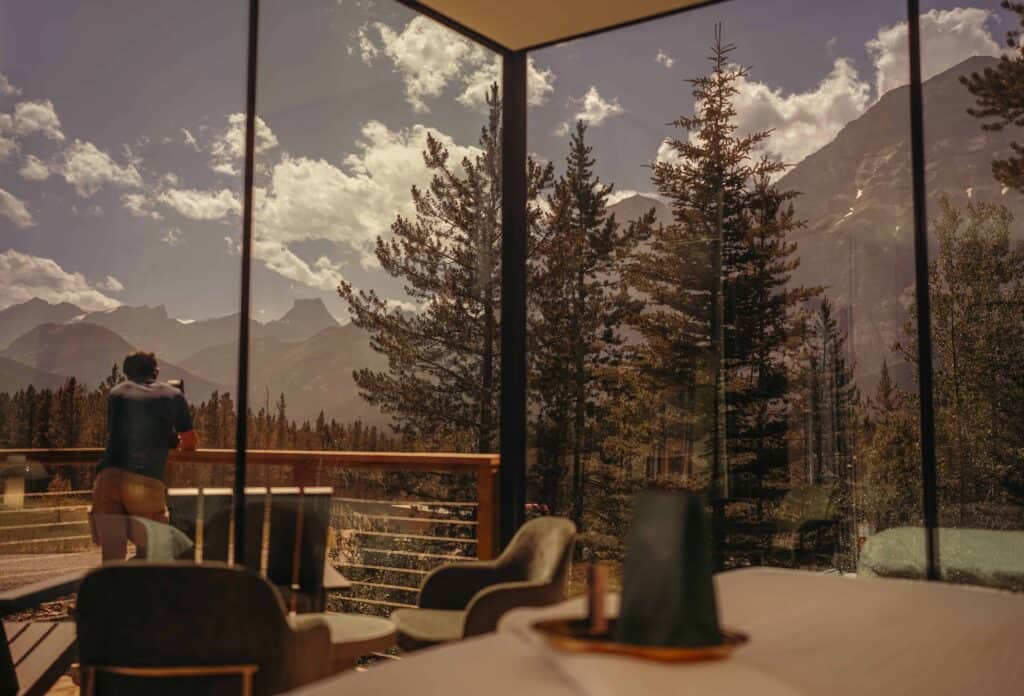 Mountain view through large glass windows at a modern mountain lodge or café, with a person enjoying the scenic landscape overlooking tall pine trees and rugged mountain peaks.