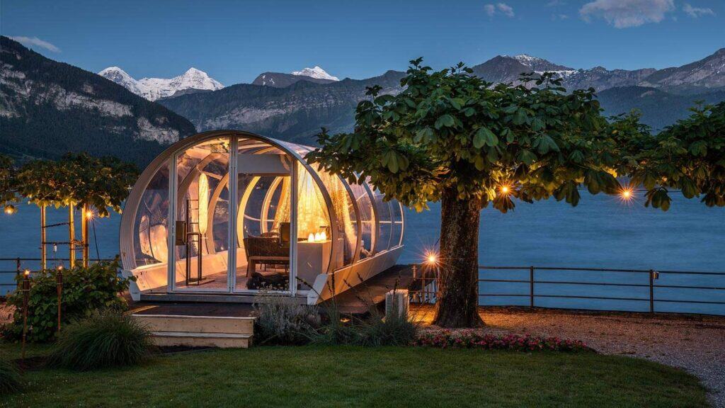 Glass honeymoon pod with scenic mountain lake view at dusk, luxurious transparent eco-structure, romantic lakeside accommodation, modern glamping experience near snow-capped peaks, serene nature retreat, outdoor leisure and hospitality, ecological lodging, waterfront outdoor hospitality featuring innovative eco-pod design.