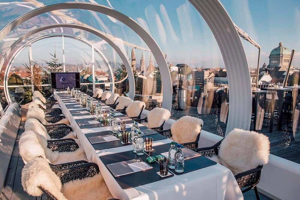 Futuristic glass enclosed rooftop dining area with elegant table setup and city skyline view, ideal for corporate events and special occasions, provided by EJH Distribution.