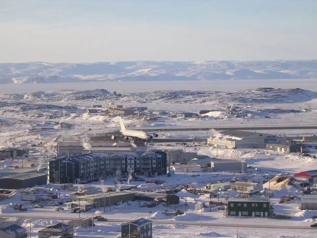 A snowy landscape of an airport and surrounding buildings in a cold climate, featuring an airplane taking off or landing on a snow-covered runway with a mountainous background.