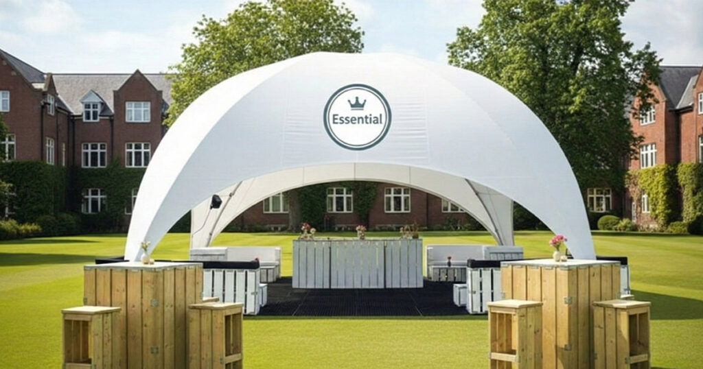 A white event tent with "Essential" branding on top, set up on a grassy lawn, featuring tables, stools, and floral accents, ideal for outdoor celebrations or gatherings in a scenic setting.