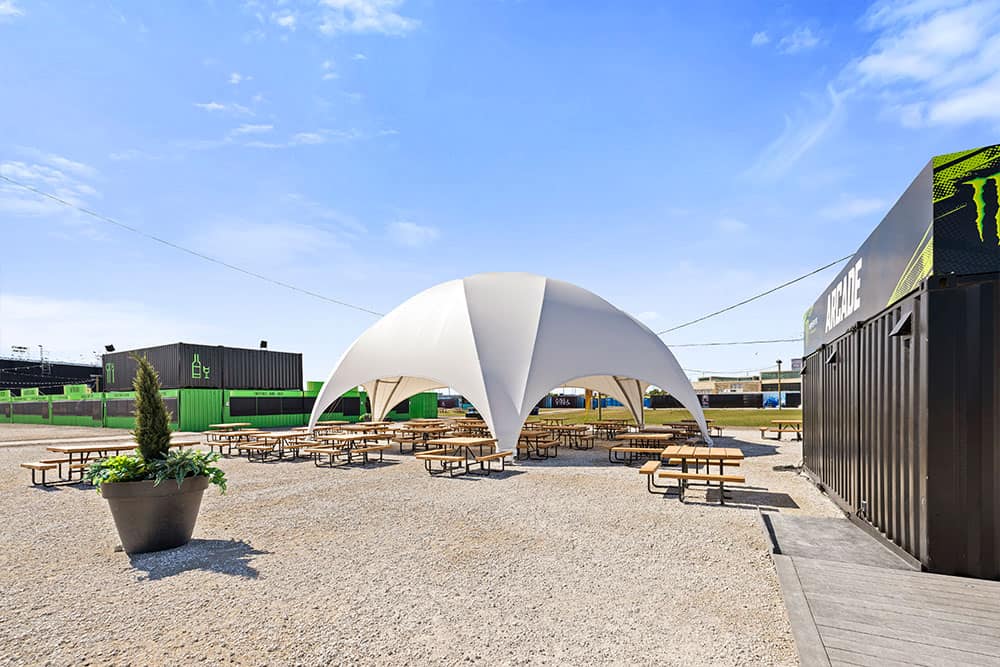 A stylish outdoor event space featuring a white modern tent, picnic tables, and shipping containers, set on a gravel surface under a clear blue sky. Ideal for outdoor gatherings, festivals, and community events, with a contemporary and functional layout.