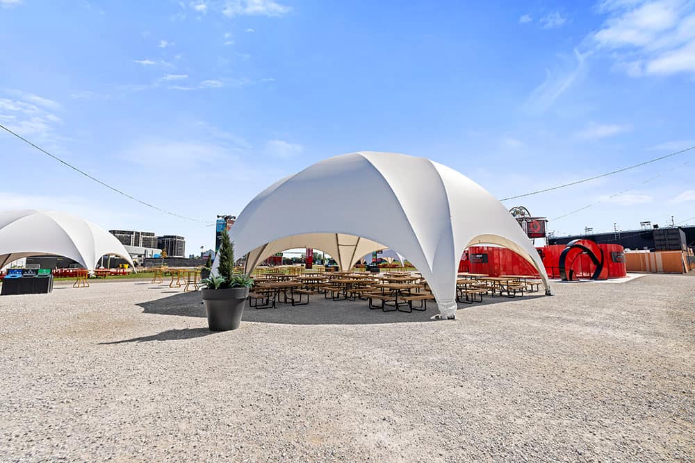 Large white canopy tents provide shaded seating areas, surrounded by gravel ground and minimalistic decor, ideal for outdoor events or festivals in urban settings.