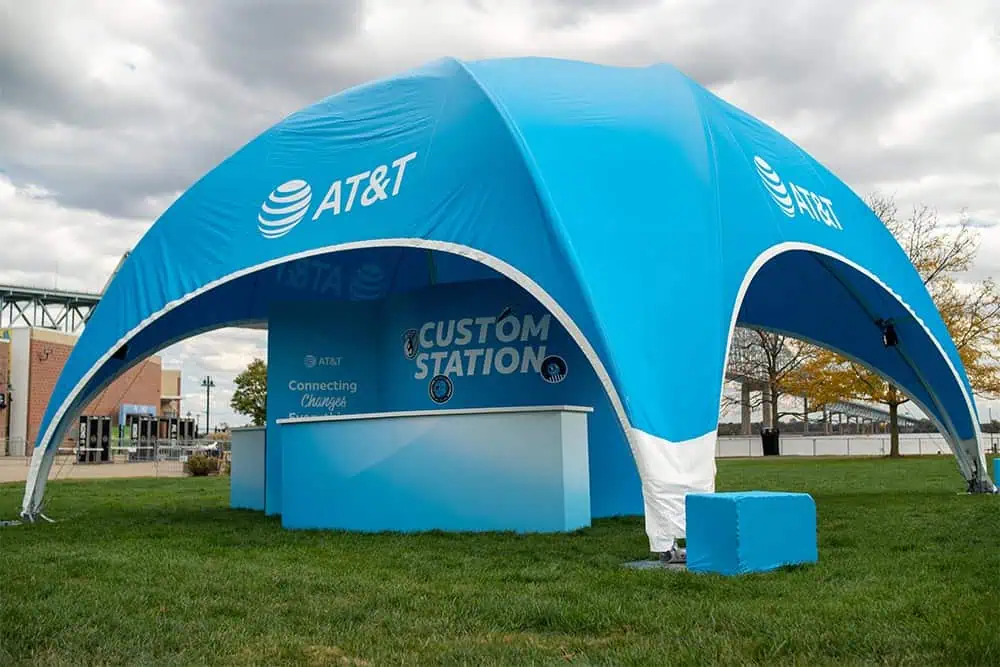 A large blue AT&T branded tent set up for customer support, featuring a "Custom Station" sign, on a grassy area with trees and a cloudy sky in the background.