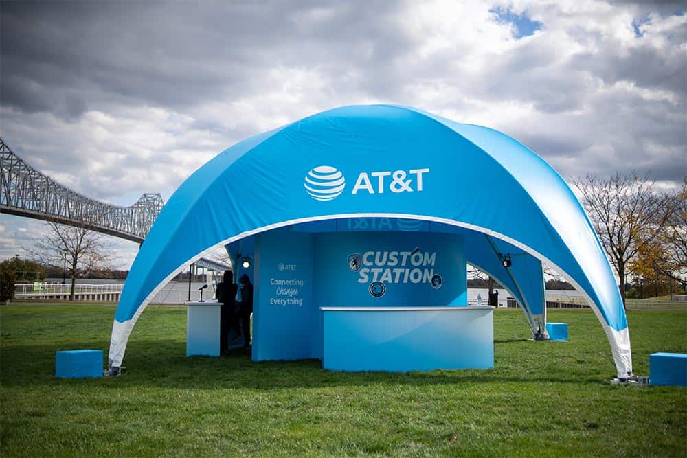 Bright blue AT&T branded canopy shelter at outdoor event with local bridge and cloudy sky background.