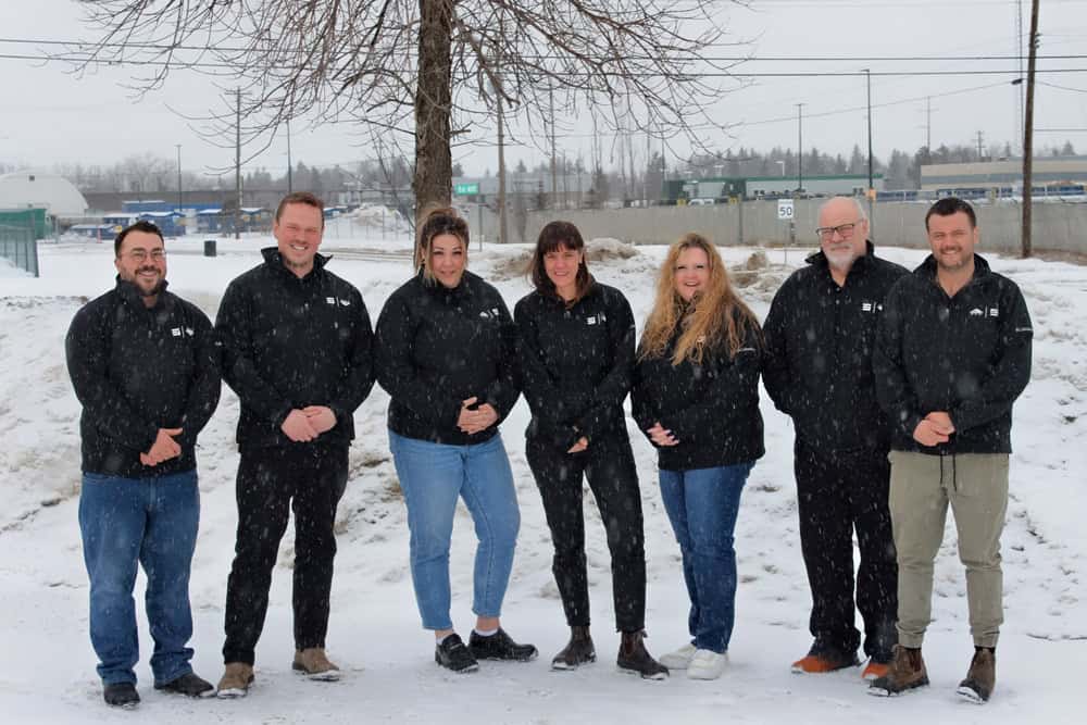 Group of six professionals dressed in black jackets, smiling and posing in a winter landscape with snow-covered ground and trees.