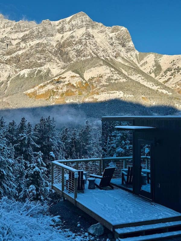 Snow-covered deck with outdoor chairs and railing, surrounded by a dense forest, against a backdrop of towering snow-capped mountains under a clear blue sky.