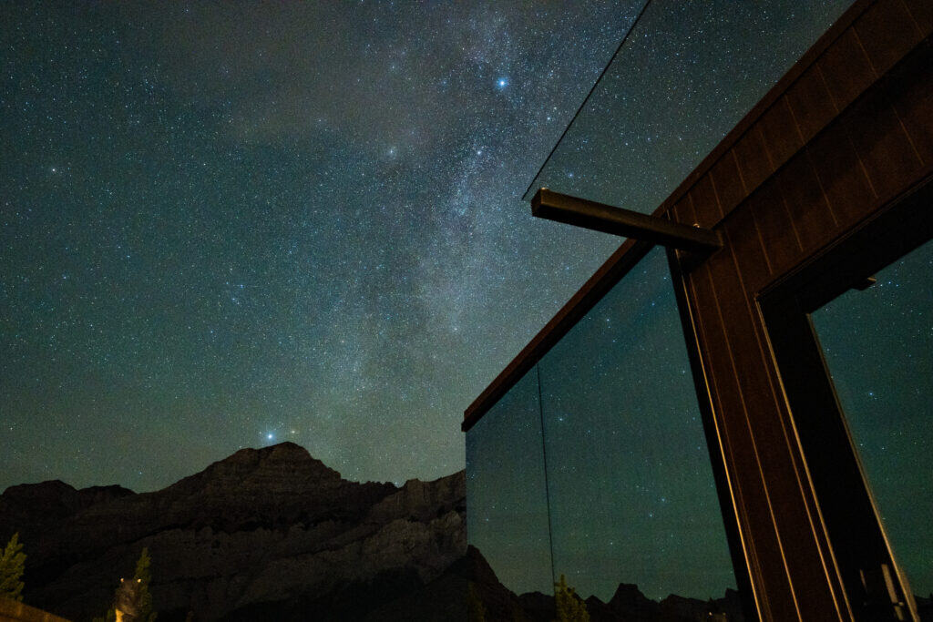 A modern tiny home with glass walls reflecting the night sky, set against a mountainous landscape under a clear, star-filled sky. Perfect for minimalist living and nature lovers.