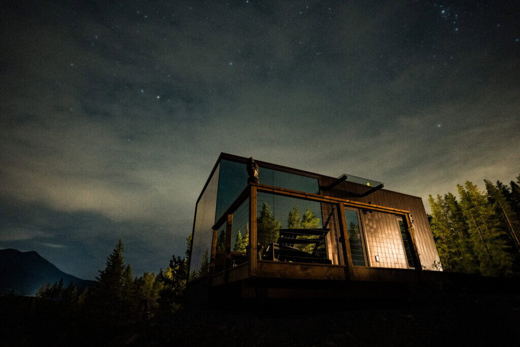 Modern tiny home with glass walls and outdoor deck, set in a forested area under a starry night sky, ideal for minimalist living and nature retreats.