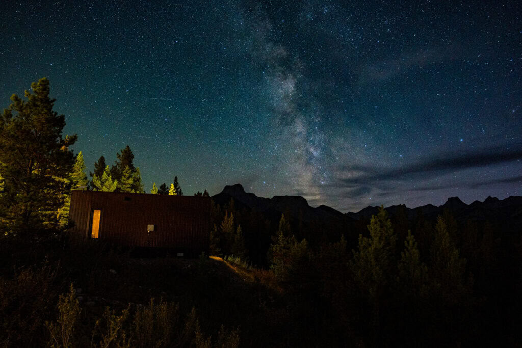 Starry night sky over a tiny home in a forested mountain area.