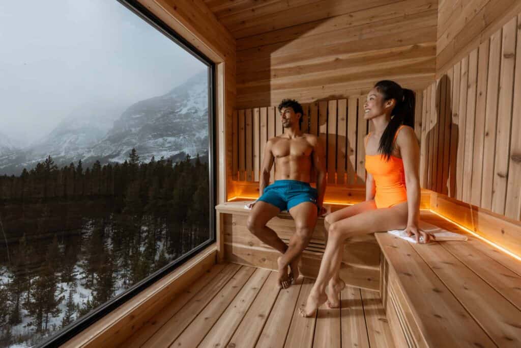 Relaxing sauna session with two people enjoying the scenic mountain landscape through a large window. The wooden interior creates a cozy and tranquil atmosphere.