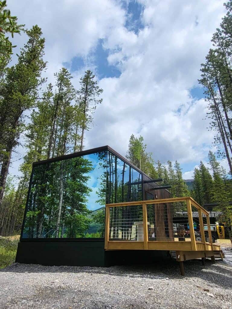 Modern glass cabin in a forest with large windows reflecting trees, surrounded by tall pine trees and a partly cloudy sky, showcasing contemporary architecture in nature.