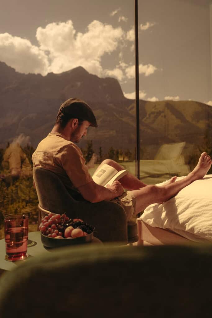 Young man relaxing inside a tiny home with large glass windows showcasing mountain scenery, reading a book, and enjoying a peaceful retreat.