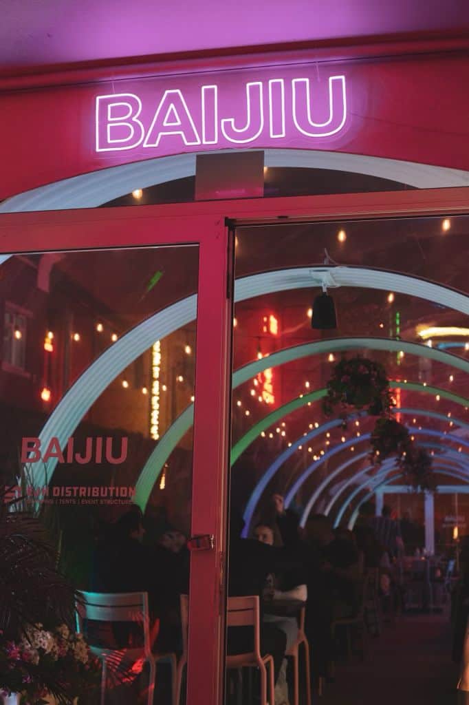 Neon-lit Baijiu sign at the entrance of a trendy Canadian patio bar, showcasing a lively atmosphere with hanging plants and illuminated arches, perfect for social gatherings.
