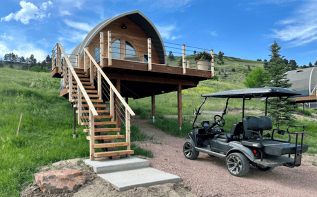 A charming A-frame cabin elevated on wooden stilts with a spacious deck, set against a lush green hillside under a bright blue sky, accompanied by a black golf cart parked nearby.