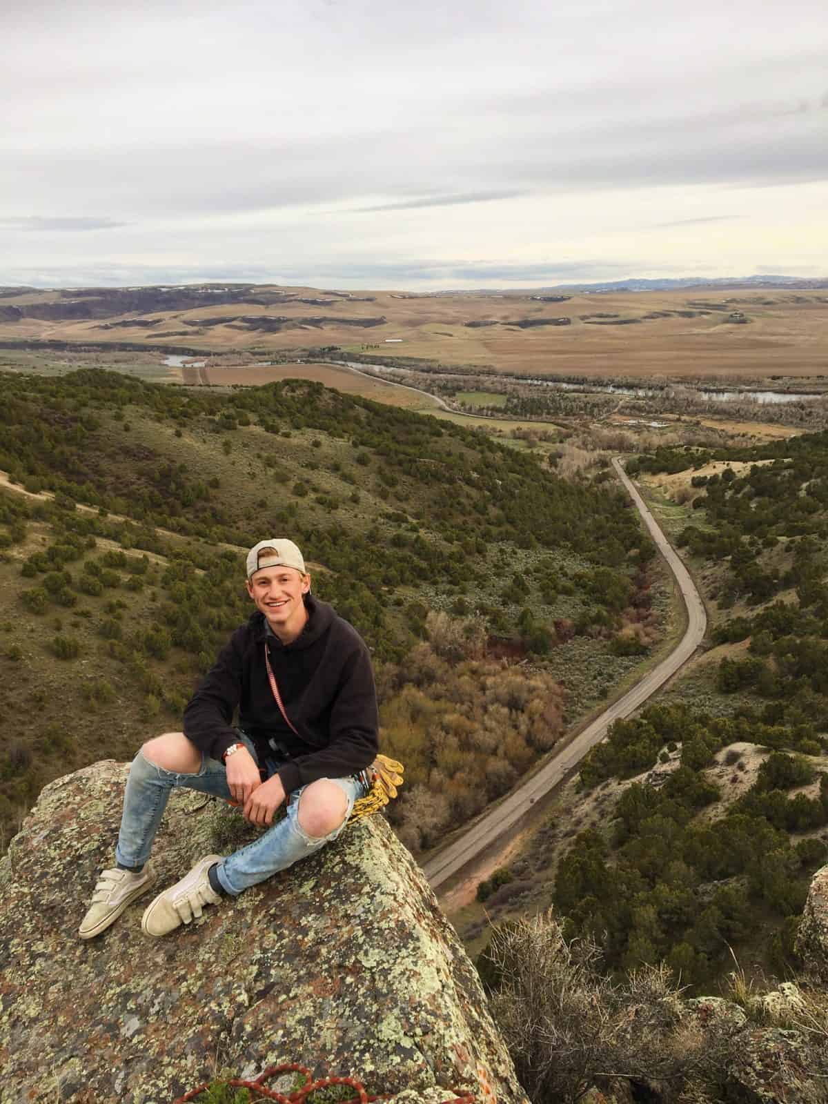 A young man in a black hoodie and ripped jeans sits on a rocky cliff overlooking a vast valley.