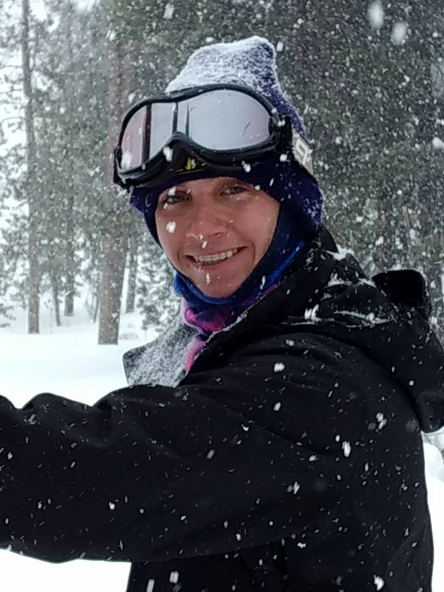 A person smiling warmly during a snowfall, dressed in winter gear including a black hooded jacket, a blue beanie, and a colorful neck gaiter. They have a pair of large ski goggles perched on their forehead, and snowflakes are visible clinging to their hat and clothing. The background shows a soft-focus, snow-covered forest with tall pine trees.