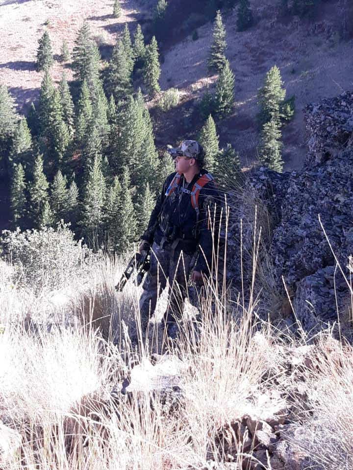 A hunter in camouflage gear stands on a rugged, grassy hillside overlooking a valley filled with evergreen trees.