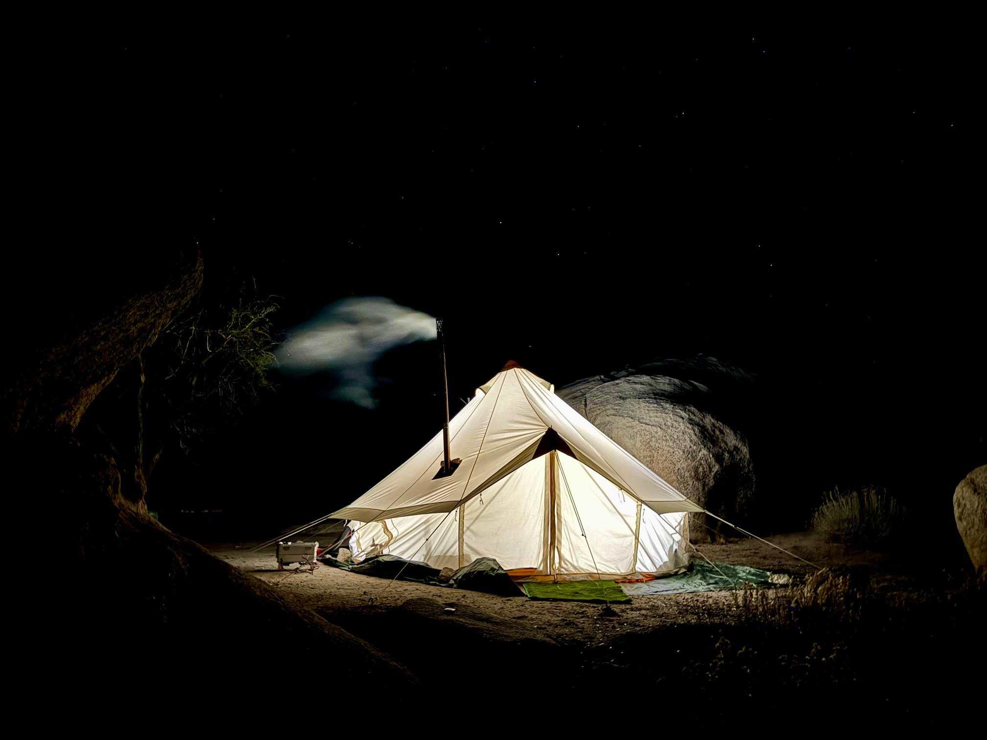 In a long-exposure night photograph, a white canvas bell tent stands out brightly against a pitch-black background, glowing with intense white light from the inside. A tall, thin metal chimney pipe rises from the side of the tent, with a soft, ghostly plume of white smoke trailing off to the left into the darkness. The tent is pitched on a dark tarp in a sandy, rugged area with a large, rounded granite boulder directly behind it and another dark, shadowed rock formation to the left. A small cooler is visible on the ground to the left of the tent. Above, a few faint stars are scattered across the dark sky.