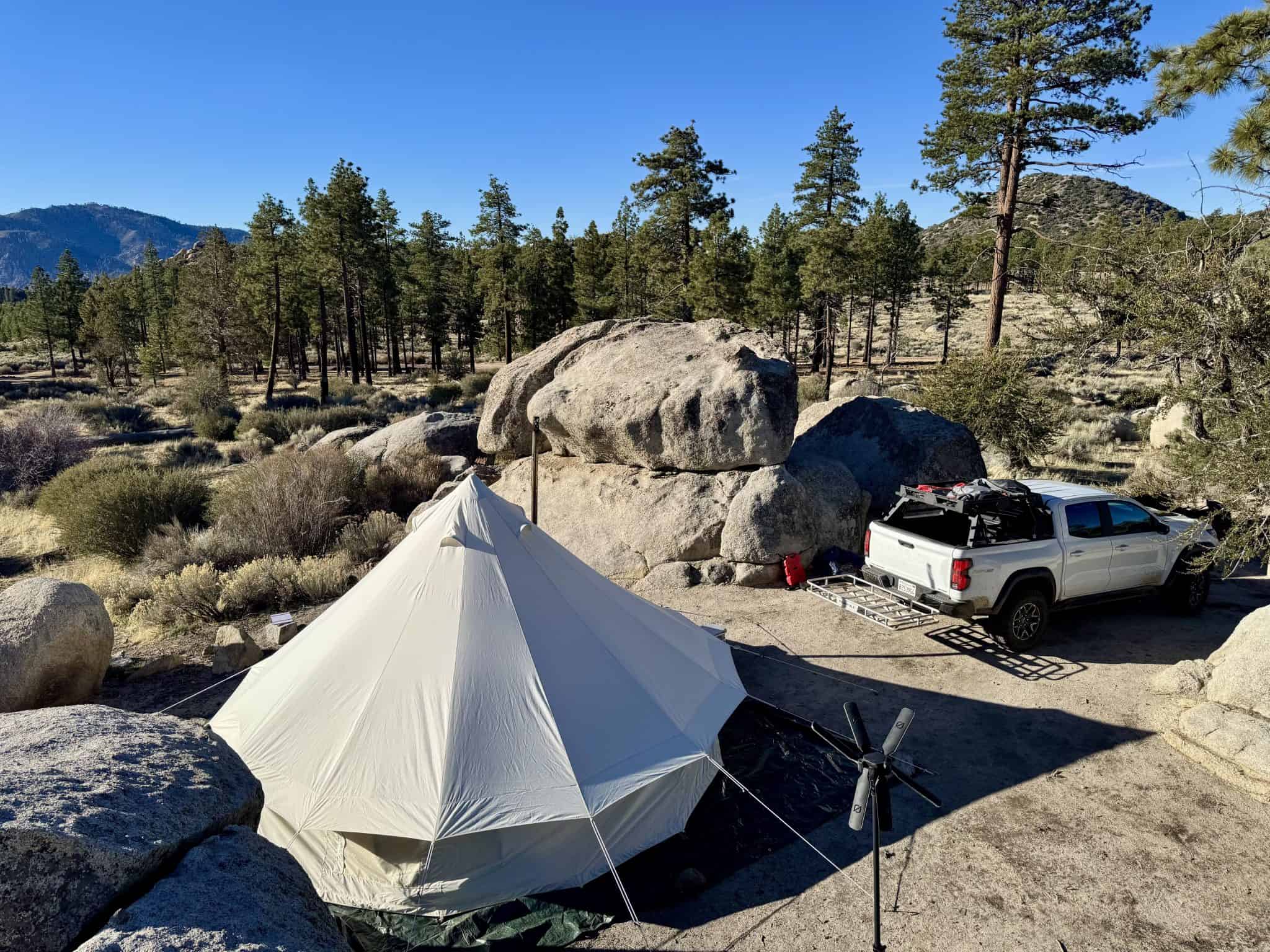 A high-angle, outdoor shot shows a large, cream-colored bell tent pitched in a rugged, high-desert landscape. Large, rounded granite boulders surround the campsite, with a white pickup truck parked nearby featuring a roof rack and gear. The background is filled with a dense forest of pine trees under a clear, bright blue sky. To the right of the tent, a black portable wind turbine stands on a tripod. The scene is bathed in bright afternoon sunlight, casting long, sharp shadows across the sandy ground.