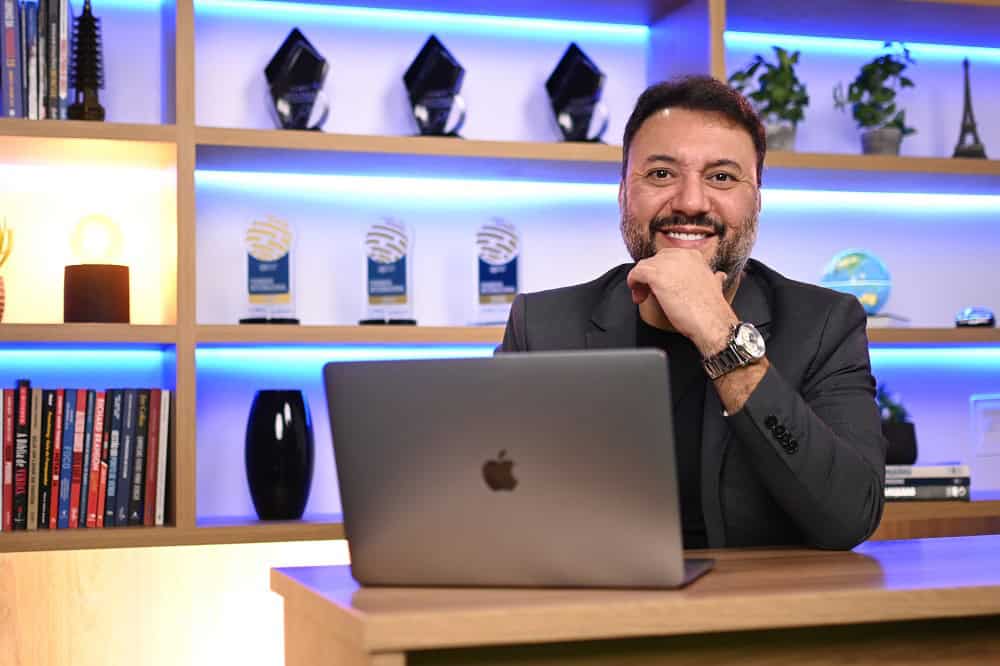 Guia do franqueado 2026 - dúvidas respondidas Henrique Mol sorridente, sentando em frente a notebook cinza. Ao fundo há prateleiras com iluminação em azul. Ilustração do texto sobre Henrique Mol.