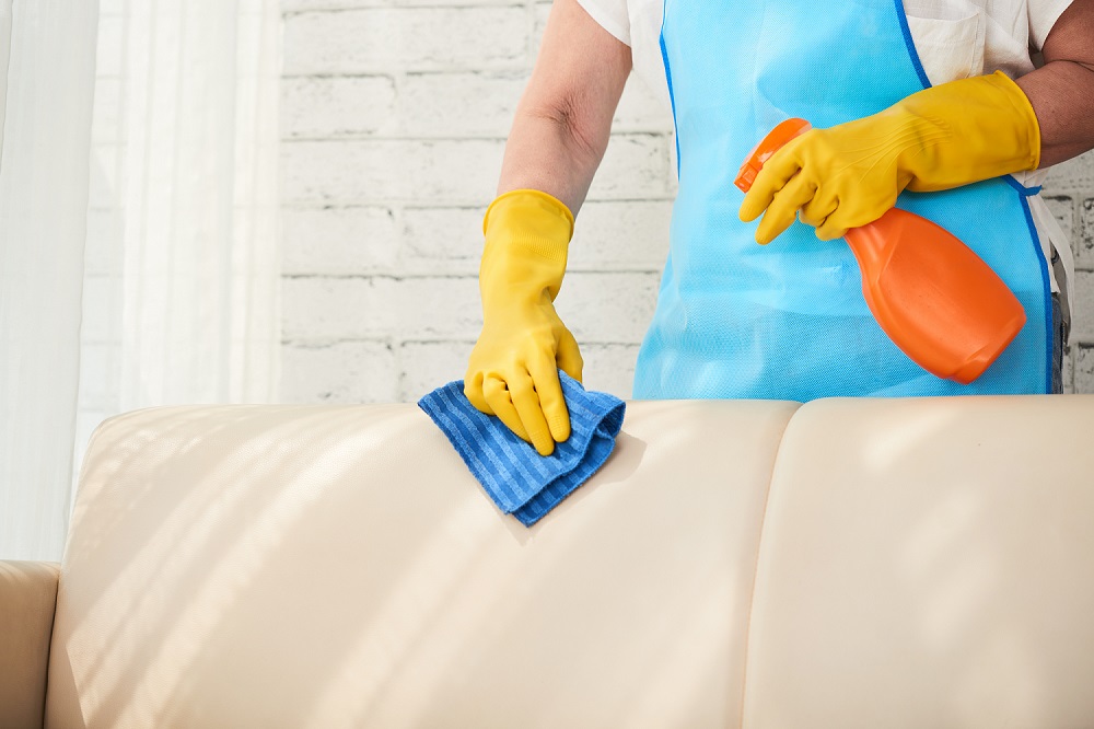 Foto de uma pessoa com avental azul e luva amarela segurando uma flanela azul e borrifador laranja para limpar sofá bege. Ao fundo temos uma parede branca. Imagem ilustrativa para texto franquia Doutor Sofá expansão
