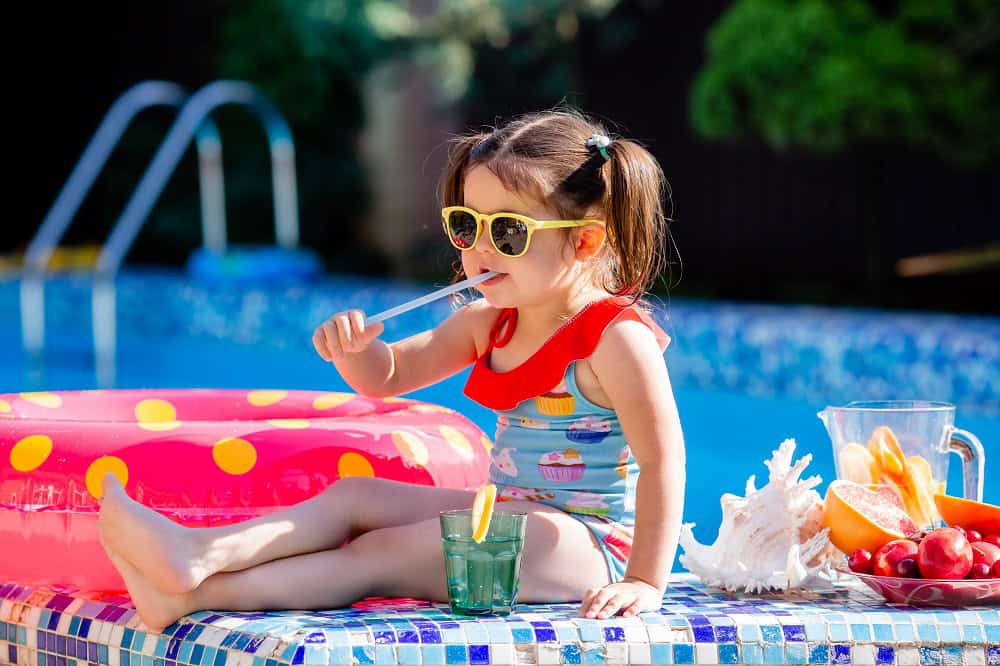 A foto traz uma criança usando maiô colorido e óculos de sol, sentada à beira da piscina. Imagem ilustrativa do texto franquia de moda infantil.