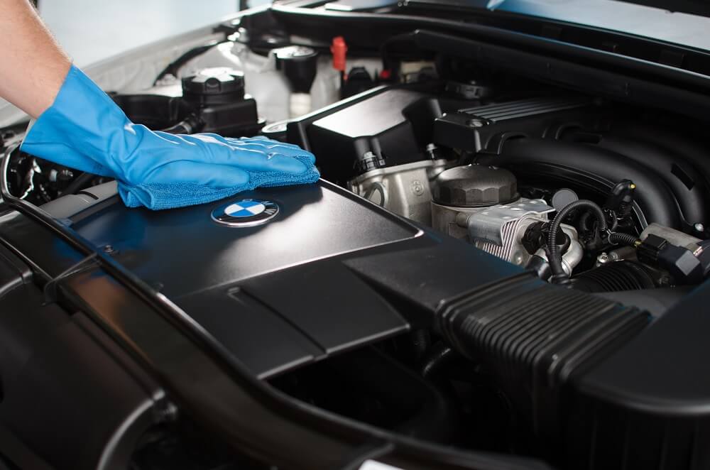 A imagem mostra a mão esquerda de uma pessoa com luva azul limpando o motor de um carro da marca BMW. Imagem ilustrativa do texto: franquia de automoveis.