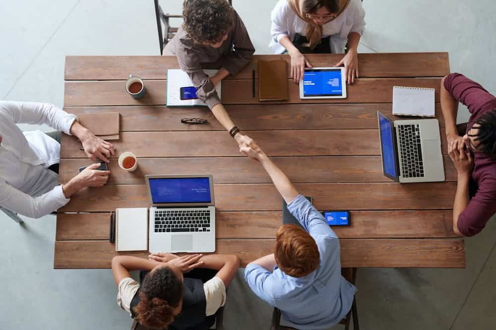 Vista superior de pessoas, sentadas em torno de mesa marrom, trabalhando com computadores. Ilustração do texto sobre modelagem de franquias.