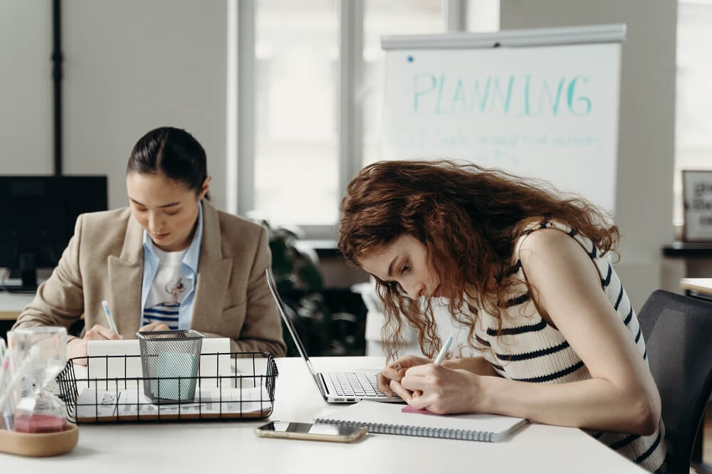 A foto traz duas mulheres trabalhando juntas, ambas fazem anotações em post-it, a jovem em primeiro plano usa blusa listrada nas cores preto e branco e a jovem em segundo plano blazer bege. Imagem ilustrativa do texto franquia para mulheres.