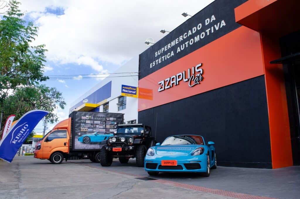 Fachada laranja de uma unidade da franquia Zaplus Car. Na frente estão estacionados um carro Porsche azul claro e um caminhão adesivado como se fossem prateleiras de produtos da Zaplus Car.
