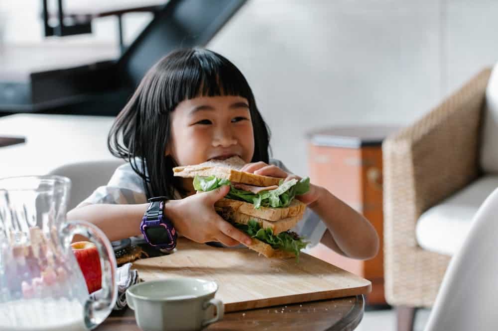 Criança sorridente, sentada à mesa, comendo um sanduíche. Ilustração do texto sobre franquias de comida.