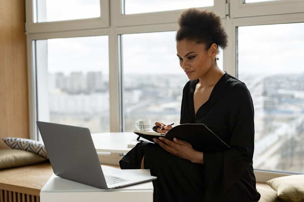 Mulher usando roupa preta, analisando o notebook, enquanto faz anotações. Ilustração do texto sobre começar o próprio negócio.
