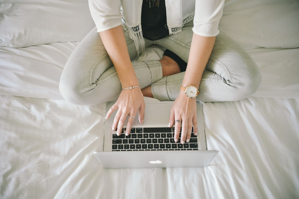 A foto traz uma mulher sentada com as pernas cruzadas em uma cama, trabalhando em seu notebook. Imagem ilustrativa do texto microfranquias baratas.